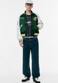 Groene en witte varsityjack met gele accenten, grijs patroon-T-shirt, losse spijkerbroek en zwart-witte schoenen. Een eenvoudige pet wordt gedragen.