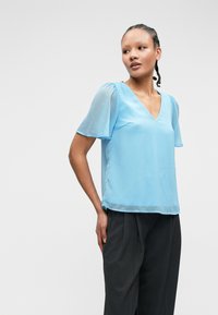 Ανοιχτό μπλε μπλούζα από μαλακό ύφασμα, με κοντά διαφανή μανίκια και V-λαιμό. Συνδυάζεται με σκούρα, κομμένα παντελόνια. Απλός και κομψός σχεδιασμός.