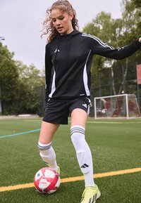 Fekete sportos kapucnis pulóver fehér oldalsávokkal, fekete rövidnadrág, fehér zoknik három fekete csík középen, és sárga focicipők, amint egy piros focilabdát cselez.