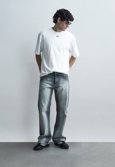 Άνδρας με σγουρά μαλλιά, που φοράει μαύρα γυαλιά ηλίου, λευκό φαρδύ μπλουζάκι, ξεθωριασμένα γκρι τζιν και μαύρα αθλητικά παπούτσια, στέκεται μπροστά σε απλή τοίχο.