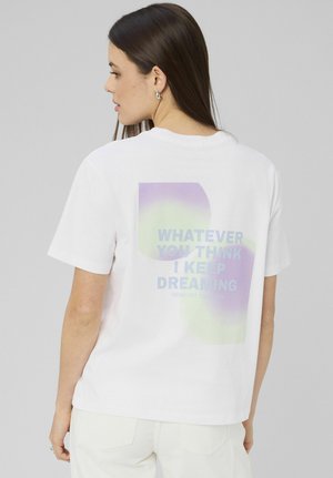 Femme aux longs cheveux bruns portant un t-shirt blanc avec un texte violet et vert au dos disant "PEU IMPORTE CE QUE TU PENSES, JE CONTINUE DE RÊVER."