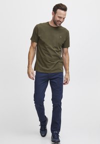 Oliva zöld pamut, rövid ujjú póló kis logóval, sötétkék, slim-fit nadrággal és fekete sportcipővel párosítva.