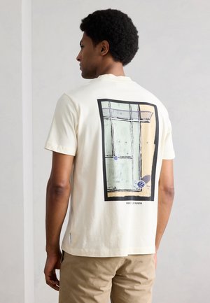 Homme aux cheveux bouclés portant un pantalon beige et un t-shirt crème avec un motif graphique rectangulaire abstrait encadré en noir dans le dos.