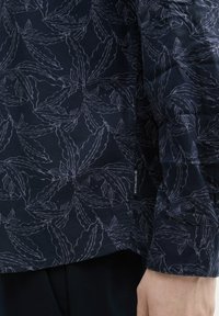 Marineblauw shirt met een botanisch bladdessin in lichtgrijs. Gemaakt van een lichte stof met een gladde textuur en een subtiel merklabel aan de zijkant.