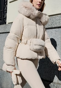 Beige vadderad jacka med huva i fuskpäls, midjebälte och sidofickor; matchande beige tajta byxor och vadderade vantar; liten mjuk midjeväska.