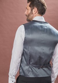 Vesta gri din satin cu un model texturat, curea din spate unică și cataramă, purtată peste o cămașă albă cu manșete cu butoni.