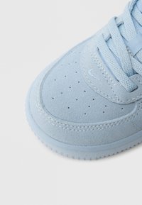 Lyseblå semsket joggesko med perforeringer på tåboksen, flate snørebånd og teksturert gummisåle med et karakteristisk mønster.