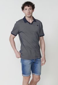 Námořnické modré a bílé vzorované polo tričko s límečkem, zkombinované se světle modrými denimovými šortkami. Tričko má na hrudi malé logo.