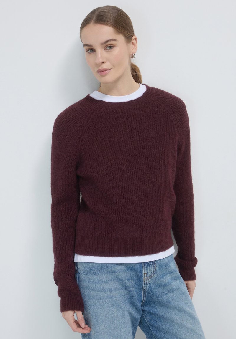 Pull en maille bordeaux avec une finition texturée, encolure ronde et manches longues, superposé sur une chemise blanche, associé à un jean bleu.