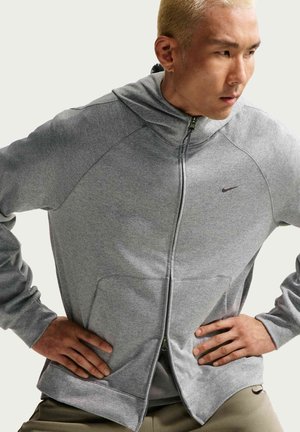 Mann mit blondem Haar, der einen grauen Kapuzenpullover mit Reißverschluss und kleinem Logo trägt, steht mit den Händen in der Hüfte und schaut nach rechts.