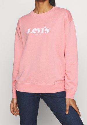 Osoba nosząca jasnoróżową bluzę Levi's z białym logo i ciemnoniebieskie dżinsy, stojąca na tle jednolitego jasnego tła.