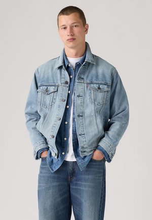 Joven de pie con las manos en los bolsillos, vistiendo chaquetas vaqueras azules claras y oscuras superpuestas sobre una camisa blanca contra un fondo gris liso.