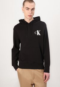 Felpa nera in cotone con tasca frontale, cappuccio con cordino e logo "CK" bianco sul petto. Presenta polsini e orlo a costine.