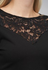 Czarny top z koronkowym dekoltem w kwiatowe wzory, w kształcie litery V, o gładkiej fakturze tkaniny.