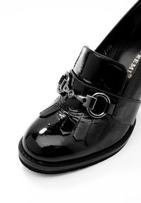 Mocassin en cuir verni noir avec franges décoratives et détail de boucle argentée sur fond blanc.