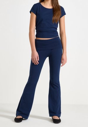 Femme portant un haut bleu marine à manches courtes et un pantalon évasé assorti avec des ballerines noires sur fond blanc.