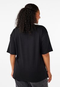 Donna dai capelli ricci che indossa una maglietta nera a maniche corte e allentata e jeans scuri, vista di spalle su uno sfondo bianco uniforme.