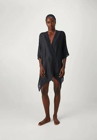 Femeie desculță îmbrăcată cu o rochie neagră lejeră, până la genunchi, cu decolteu în V, stând pe un fundal deschis simplu.