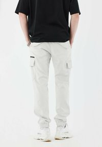 Lichtgrijze cargobroek met zijzakken, elastische boorden en een relaxed fit, gecombineerd met witte sneakers tegen een effen achtergrond.