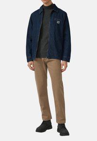 Jeansjacke in Dunkelblau mit Reißverschluss, Brusttasche, getragen über einem dunkelgrauen Pullover, kombiniert mit beigen Hosen und schwarzen Stiefeln.