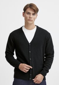 Svart ullcardigan med V-hals, ribbestrikkede mansjetter og frontlukking med knapper. Har en myk tekstur og klassisk design. Brukes over en hvit skjorte.