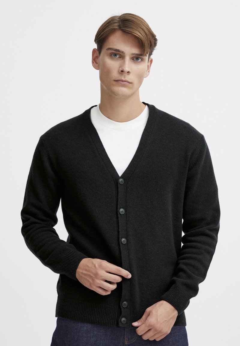 Svart ullcardigan med V-hals, ribbestrikkede mansjetter og frontlukking med knapper. Har en myk tekstur og klassisk design. Brukes over en hvit skjorte.