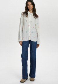 Camicia bianca a maniche lunghe con bottoni e ricami floreali, abbinata a pantaloni in denim blu a palazzo e stivaletti beige. Tessuto morbido, vestibilità comoda.