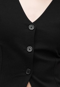 Μαύρο κασμίρε ζακέτα με V λαιμόκοψη και τρία μαύρα κουμπιά που κλείνουν μπροστά, φορεμένο από άτομο με ορατό πάνω μέρος του στήθους και λαιμό.