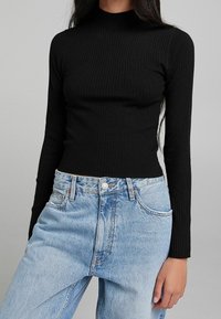 Haut noir côtelé à manches longues avec col montant, associé à un jean bleu clair taille haute à coupe classique et texture délavée.