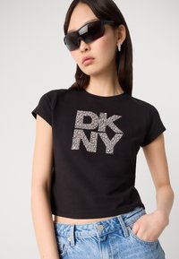 Schwarzes, kurzes T-Shirt mit einem strukturierten silbernen Strass-„DKNY“-Grafik. Kurze Ärmel und nahtloser Ausschnitt. Kombiniert mit blauen Jeansshorts.