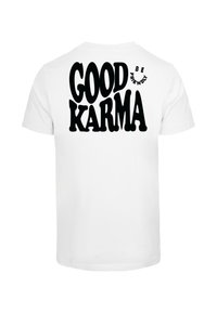 Bílá bavlněná trička s velkým černým grafickým textem na zádech, který zní "DOBRÁ KARMA" a menším textem "BUĎ PŘÁTELSKÝ", zaoblené okraje.