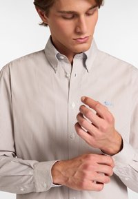 Camisa de botones en color beige claro con finas rayas verticales. Presenta un cuello puntiagudo y pequeño logo azul en el pecho. Tejido de algodón.