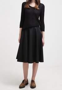 Vrouw in een zwarte gestructureerde langslavend top, zwarte midi-rok en leren veterschoenen met luipaardprint, staande tegen een effen achtergrond.