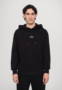 Black hoodie made of soft fabric, featuring a front pocket, drawstring hood, and a small gold logo on the chest. Fitted at the waist and cuffs.