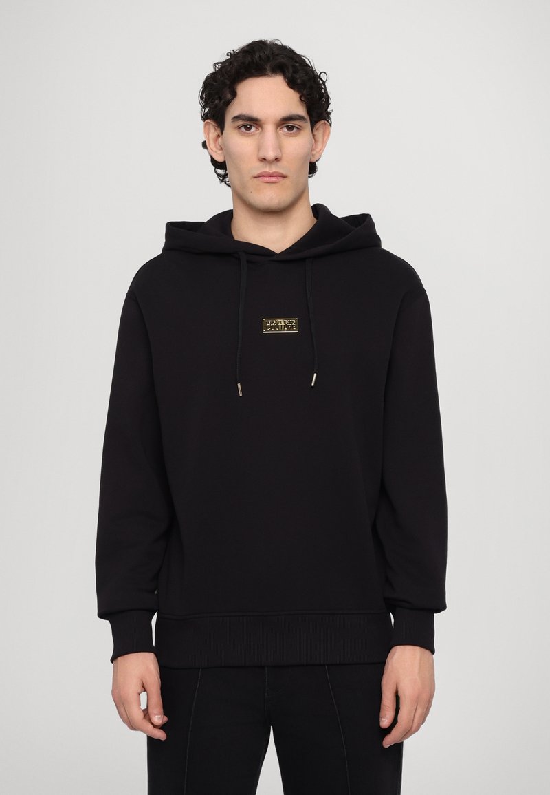 Black hoodie made of soft fabric, featuring a front pocket, drawstring hood, and a small gold logo on the chest. Fitted at the waist and cuffs.