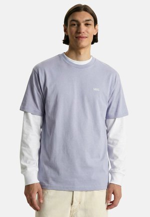 Giovane uomo che indossa una maglietta Vans color lavanda a maniche corte sopra una camicia bianca a maniche lunghe e pantaloni beige, sorridente con le mani rilassate.
