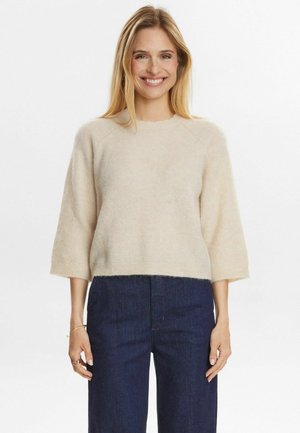 Beige, fluffy trui met een ronde halslijn en driekwartmouwen, met een cropped zoom en zachte textuur. Gecombineerd met donkere spijkerbroek.