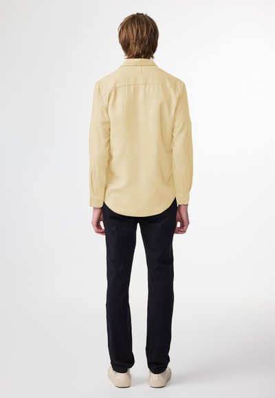 Helles, gelbes Langarmshirt mit Knopfdetails, Rückenschößchen und abgerundetem Saum; kombiniert mit schwarzen Hosen und hellen Schuhen.