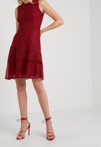 Robe en dentelle rouge avec un corsage ajusté, design sans manches, jupe à volants et ourlet festonné. Associée à des sandales à talons hauts rouges à lanières.