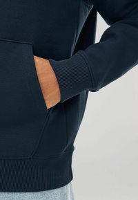 Sudadera azul marino con puños de canalé, que cuenta con un bolsillo tipo canguro y una textura suave. Parte de la manga visible con una mano dentro del bolsillo.