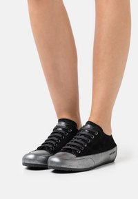 Ben som bär svarta och silverfärgade låga sneakers med snörning, stående på en vit yta.