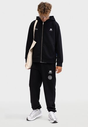 Jeune personne portant un sweat à capuche noir et un pantalon de survêtement avec des logos blancs, des lunettes transparentes, des baskets blanches, et portant un sac à bandoulière beige, regardant vers le bas.