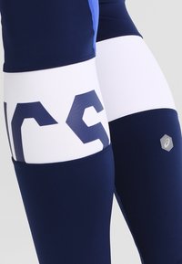 Granatowe i białe legginsy sportowe z geometrycznym wzorem, z wyraźnym logo w ciemnym granacie oraz odblaskowym akcentem na dolnej części nogi.