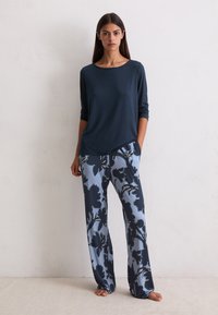 Donna con capelli lunghi e scuri, in piedi a piedi nudi contro un muro bianco, indossa un top blu navy a maniche lunghe e pantaloni larghi azzurri chiari con una stampa floreale scura.