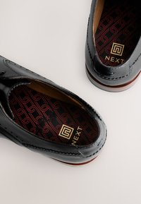 Black leather shoes feature a smooth finish, stitched detailing, and a brown sole. The interior has a patterned red and black insole with gold branding.