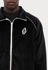 La veste de survêtement en velours noir présente des garnitures blanches et une fermeture éclair. Un logo notable en forme de cône de glace blanc est visible sur la partie gauche de la poitrine.