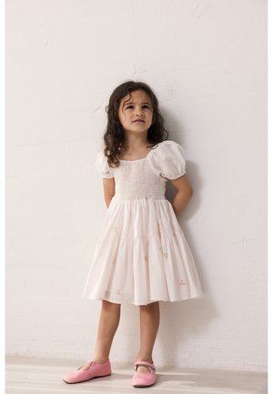 Νεαρή κοπέλα με κατσαρά μαλλιά, φορώντας λευκό φόρεμα με φουσκωτά μανίκια και ροζ παπούτσια, που στέκεται μπροστά σε ένα απλό λευκό τοίχο.