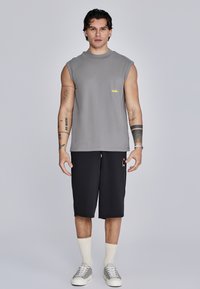 Tricou gri fără mâneci cu logo galben, șorturi negre până la genunchi, adidași albi și gri și șosete de culoare crem. Fundal simplu.