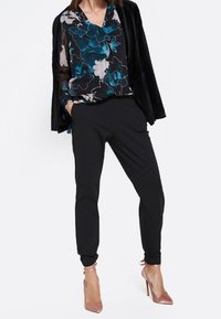 Schwarze Hosen mit schmalem Schnitt, kombiniert mit einer durchsichtigen schwarzen Bluse mit blau-weißer Blumenmuster. Abgerundet mit nudefarbenen High Heels.