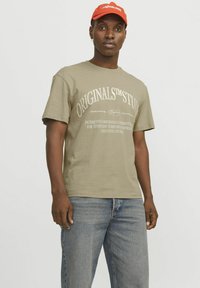 Olijfgroene katoenen t-shirt met ronde hals, korte mouwen en witte bedrukte tekst. Gekoppeld aan lichtblauwe denim jeans.